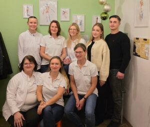 Acht Personen lächeln freundlich in weißer und beiger Kleidung vor grüner Wand