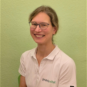 Eine lächelnde Frau mit Brille und lockigem Haar trägt ein weißes Polo vor grüner Wand